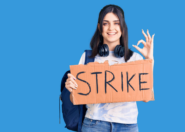 Joven hermosa chica con mochila de estudiante sosteniendo bandera de huelga haciendo signo de ok con los dedos, sonriendo gesto amistoso excelente símbolo  - Foto, Imagen