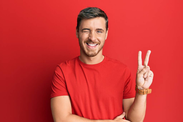 Handsome caucasian man wearing casual red tshirt smiling with happy face winking at the camera doing victory sign with fingers. number two.  - Photo, Image