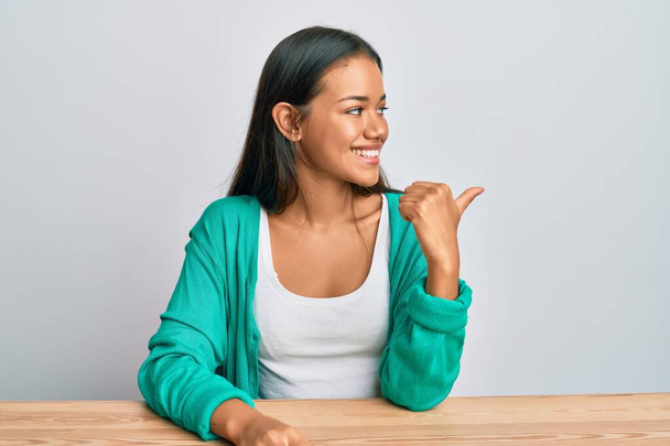 Mulher hispânica bonita vestindo roupas casuais sentado na mesa apontando polegar até o lado sorrindo feliz com a boca aberta  - Foto, Imagem