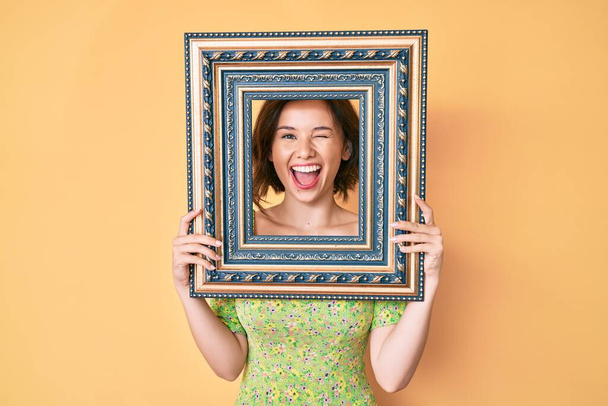 Young beautiful woman holding empty frame winking looking at the camera with sexy expression, cheerful and happy face.  - Photo, Image