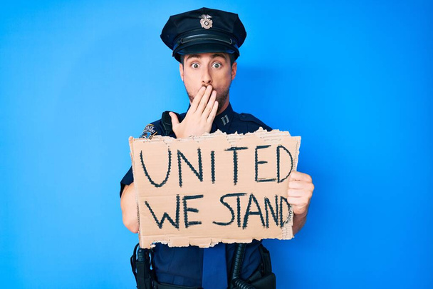 Jonge blanke man in politie-uniform verenigd staan we met een spandoek over mond met de hand, geschokt en bang voor fouten. verraste uitdrukking  - Foto, afbeelding