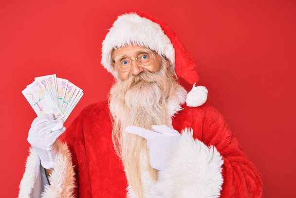 Staruszek z siwymi włosami i długą brodą w kostiumie Świętego Mikołaja trzymający kolumbijskie peso uśmiechnięty szczęśliwy ze wskazaniem ręką i palcem  - Zdjęcie, obraz
