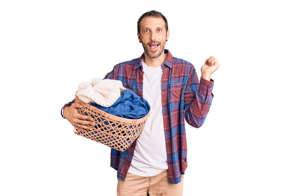 Young handsome man holding laundry basket screaming proud, celebrating victory and success very excited with raised arms  - Photo, Image