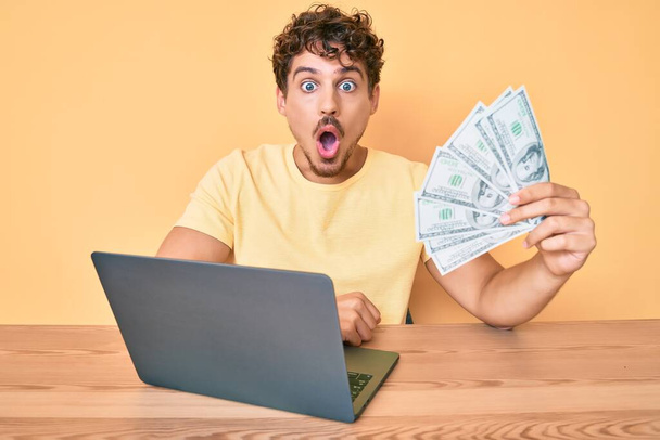 młody biały człowiek z kręconymi włosami działa za pomocą laptopa komputerowego trzymając usa dolarów banknotów przerażony i zdumiony z otwartymi ustami dla zaskoczenia, niedowierzanie twarz  - Zdjęcie, obraz