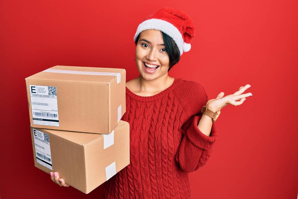 Gyönyörű fiatal nő rövid haj visel karácsonyi kalap gazdaság szállítási csomagok ünneplő győzelem boldog mosollyal és a győztes kifejezés emelt kézzel  - Fotó, kép