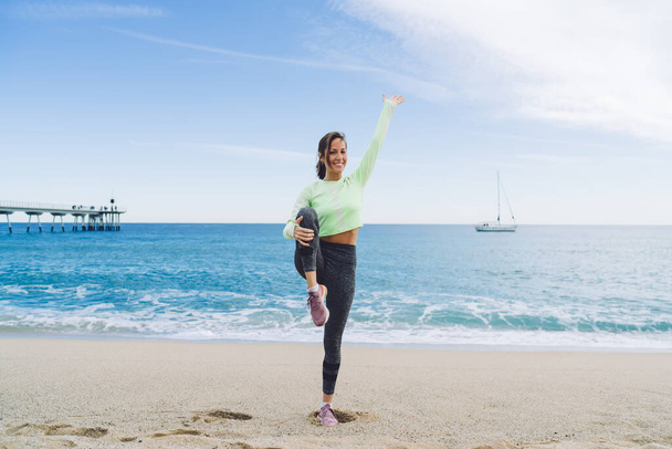 Full length portrait of Caucasian woman dressed in tracksuit stretching legs posing at seashore coastline, slim fit girl looking at camera keeping flexible body shape and healthy lifestyle - Foto, Bild
