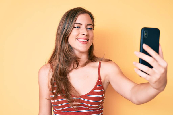 Beautiful young caucasian woman taking a selfie photo with smartphone looking positive and happy standing and smiling with a confident smile showing teeth  - Foto, afbeelding