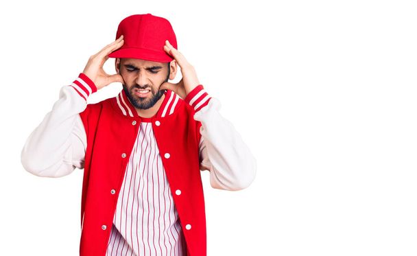 Young handsome man with beard wearing baseball jacket and cap suffering from headache desperate and stressed because pain and migraine. hands on head.  - Фото, изображение