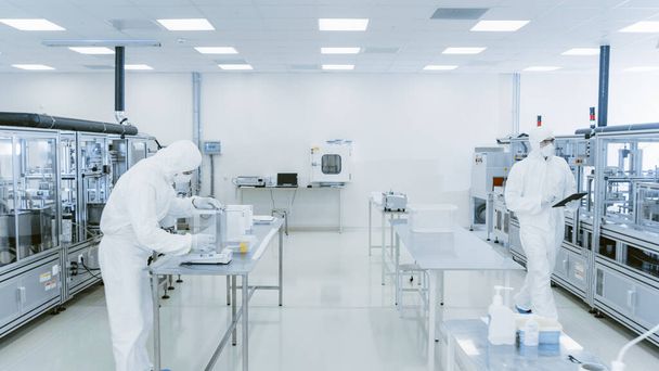 Quality Control Check: Scientist Using Digital Tablet Computer and wearing Protective Suit walks through Manufacturing Laboratory. Product Manufacturing: Pharmaceutics, Semiconductors, Biotechnology. - Photo, Image