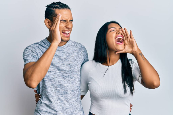 Mladý latinskoamerický pár, oblečený do neformálního oblečení, křičící a křičící na stranu s rukou na ústech. komunikační koncept.  - Fotografie, Obrázek