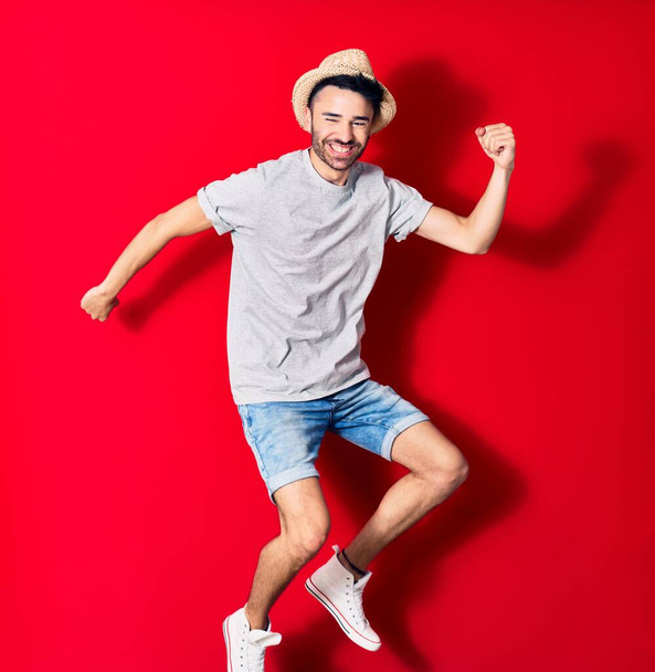 Young handsome hispanic man wearing casual clothes and summer hat smiling happy. Jumping with smile on face celebrating with fists up over isolated red background. - Photo, Image