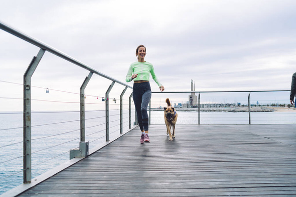 Low angle of active adult female in sporty outfit smiling and running with adorable obedient dog on wooden pier while training against cloudy sky and cityscape - Фото, зображення