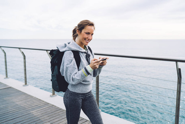Half length portrait of Caucasian woman with modern smartphone technology in hands smiling at camera, cheerful female tourist with backpack connecting to roaming internet for mobility messaging - Foto, Imagem