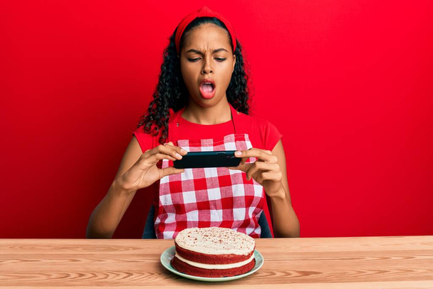 Giovane ragazza afroamericana indossando grembiule panettiere fare immagine torta di carote in faccia shock, cercando scettico e sarcastico, sorpreso con la bocca aperta  - Foto, immagini