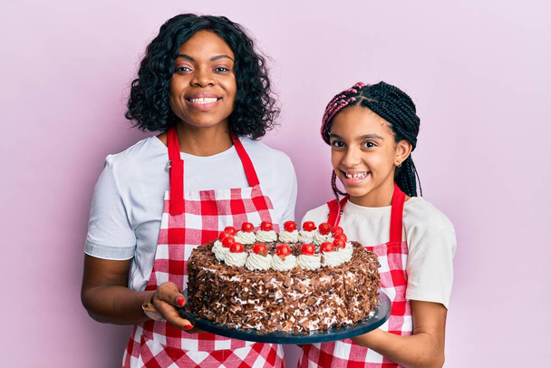 美しいアフリカ系アメリカ人の母親と娘は、パン屋のエプロンを身に着けて、笑顔で笑顔で自家製ケーキを開催します。歯を見せて.  - 写真・画像