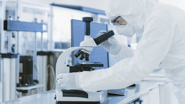 In Laboratory Scientist in Protective Clothes Doing Research Uses Microscope and Personal Computer. Modern Manufactory Producing Semiconductors and Pharmaceutical Items. - 写真・画像