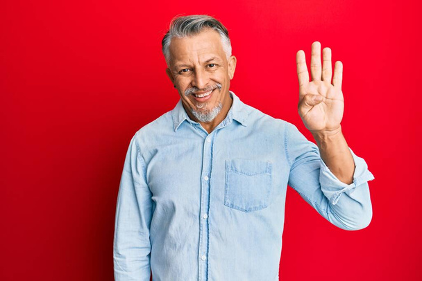 Grauhaariger Mann mittleren Alters in lässiger Kleidung, der mit Finger Nummer vier zeigt und nach oben zeigt, während er selbstbewusst und glücklich lächelt.  - Foto, Bild