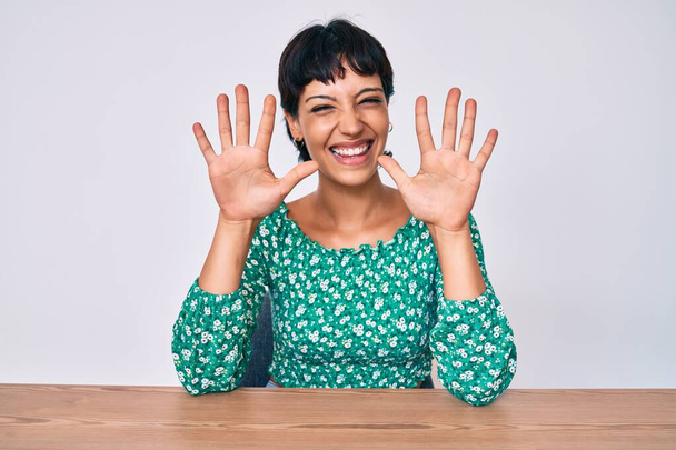 Schöne brünette Frau in lässiger Kleidung sitzt auf dem Tisch und zeigt mit Finger Nummer zehn nach oben, während sie selbstbewusst und glücklich lächelt.  - Foto, Bild
