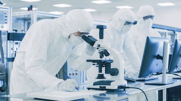 In Laboratory Scientists in Protective Clothes Doing Research, Using Microscope and Writing Data to a Notebook. Modern Manufactory Producing Semiconductors and Pharmaceutical Items. - Photo, image