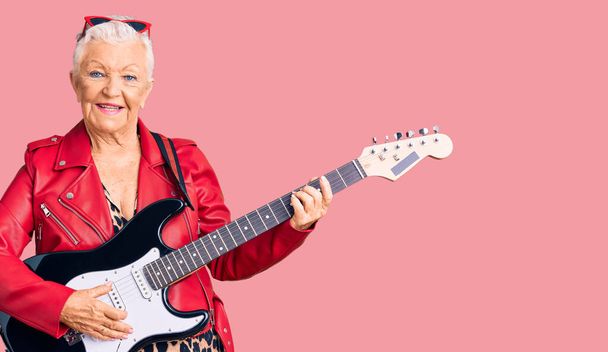 Senior hermosa mujer con ojos azules y cabello gris con un aspecto moderno tocando la guitarra eléctrica con una sonrisa feliz y fresca en la cara. persona afortunada.  - Foto, imagen