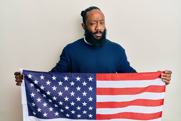 Junger afrikanisch-amerikanischer Mann, der die Flagge der Vereinigten Staaten in der Hand hält, ist skeptisch und nervös und runzelt ob des Problems die Stirn. negative person.  - Foto, Bild