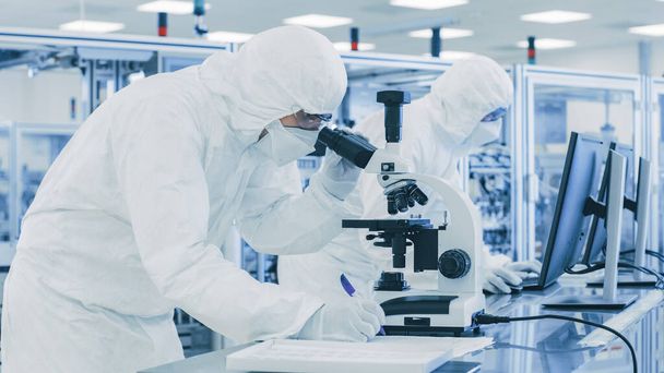 In Laboratory Scientist in Protective Clothes Doing Research, Using Microscope and Writing Down Data. Workers Working on a Modern Manufactory Producing Semiconductors and Pharmaceutical Items. - Фото, зображення