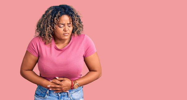 Jonge Afro-Amerikaanse plus size vrouw draagt casual kleding met de hand op de maag omdat indigestie, pijnlijke ziekte zich onwel voelt. pijnconcept.  - Foto, afbeelding