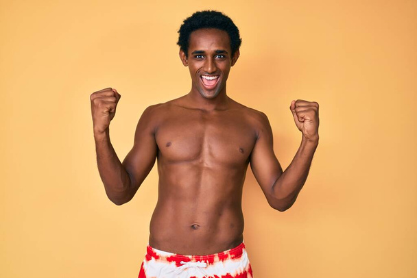Afrikai jóképű férfi fürdőruhát visel sikoltozó büszke, ünneplő győzelem és siker nagyon izgatott felemelt karokkal  - Fotó, kép