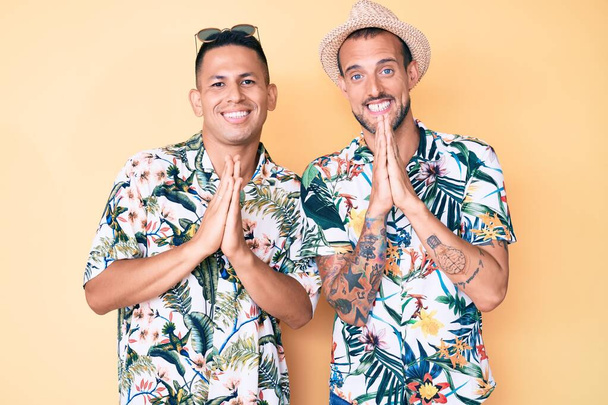 Joven pareja gay de dos hombres con sombrero de verano y camisa hawaiana rezando con las manos juntas pidiendo perdón sonriendo confiado.  - Foto, Imagen