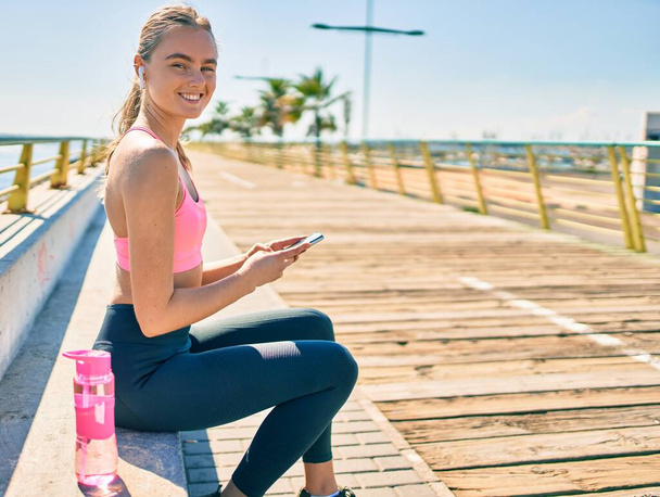 Joven deportista rubia haciendo ejercicio usando smartphone sentado en el banco en el paseo marítimo. - Foto, imagen