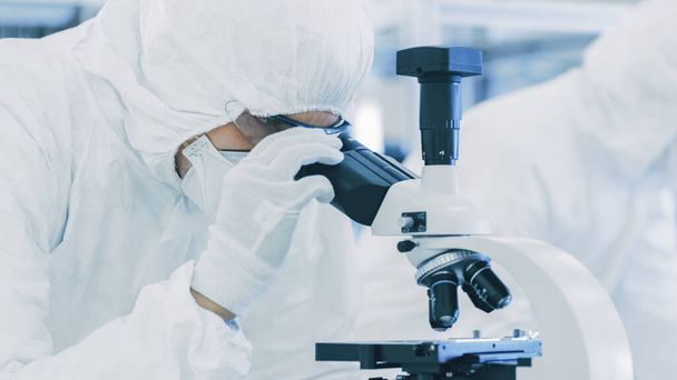 In Laboratory Scientist in Protective Clothes Doing Research, Using Microscope and Writing Down Data. Workers Working on a Modern Manufactory Producing Semiconductors and Pharmaceutical Items. - Фото, изображение