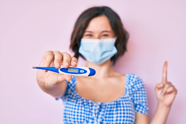 Young beautiful woman wearing medical mask holding holding thermometer surprised with an idea or question pointing finger with happy face, number one  - Photo, Image