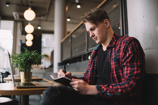 alulról fókuszált fiatal férfi alkalmi ruhák jegyzetel notebook, miközben ül az ebédlőben laptop és csinál távoli munkát - Fotó, kép