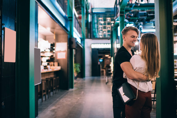 Smiling young man and woman in casual clothes hugging each other while standing near pillar in modern cafe while having meeting - Foto, Imagen