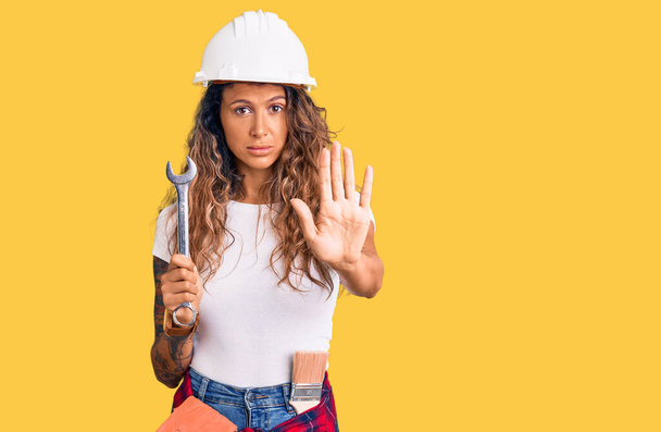 Jonge Latijns-Amerikaanse vrouw met tatoeage dragen van harde hoed en bouwer kleding houden moersleutel met open hand doen stopteken met ernstige en zelfverzekerde uitdrukking, verdediging gebaar  - Foto, afbeelding