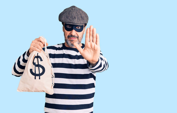 Middle age handsome man wearing burglar mask holding money bag with open hand doing stop sign with serious and confident expression, defense gesture  - Photo, Image