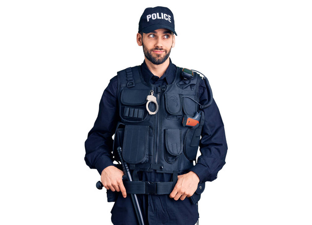 Νεαρός όμορφος άντρας με γενειάδα φορώντας αστυνομική στολή χαμογελώντας κοιτώντας στο πλάι και κοιτάζοντας αλλού σκεπτόμενος.  - Φωτογραφία, εικόνα