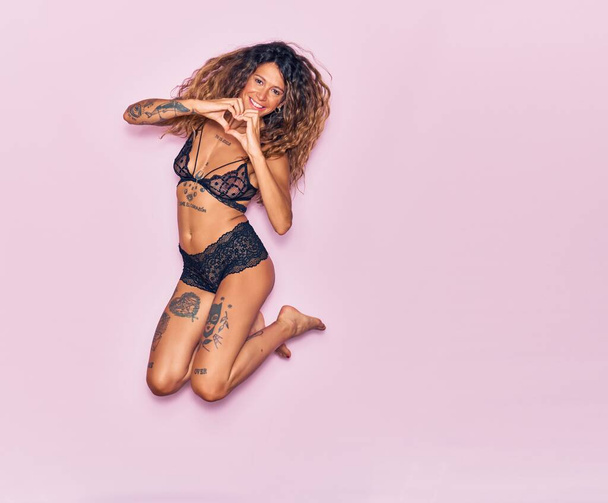 Young beautiful woman with tattoo wearing lingerie smiling happy. Jumping with smile on face doing heart shape with hands over isolated pink background - Photo, Image