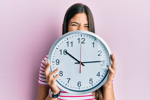 młoda brunetka kobieta trzyma duży zegar zakrywając twarz wystający język szczęśliwy z zabawnym wyrazem.  - Zdjęcie, obraz