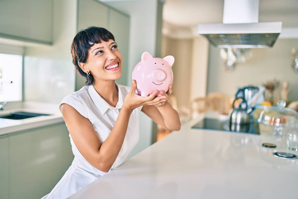 Junge brünette Frau lächelt glücklich und zeigt stolzes Sparschwein mit Ersparnissen - Foto, Bild