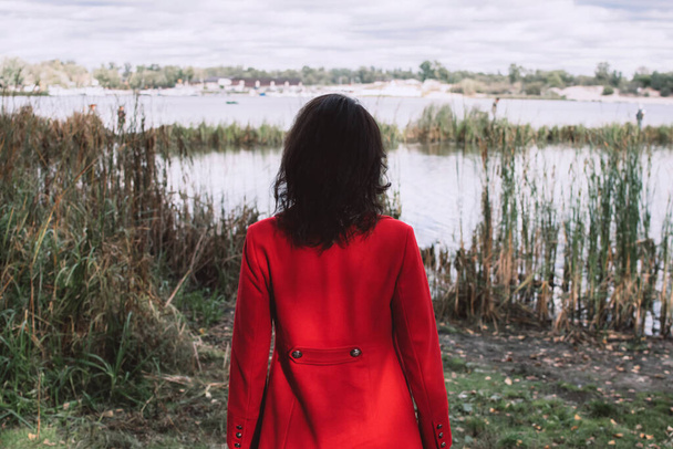 Woman wearing red coat backwards and looking river or lake landscape outdoors. Fall concept. - Photo, Image