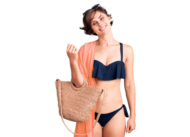 Beautiful young woman with short hair wearing bikini and hat holding summer wicker handbag looking positive and happy standing and smiling with a confident smile showing teeth  - Fotoğraf, Görsel