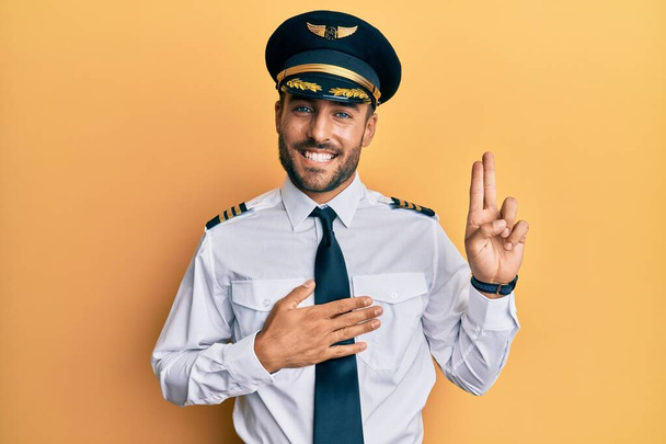 Bonito homem hispânico vestindo uniforme piloto de avião sorrindo jurando com a mão no peito e dedos para cima, fazendo uma promessa de lealdade juramento  - Foto, Imagem