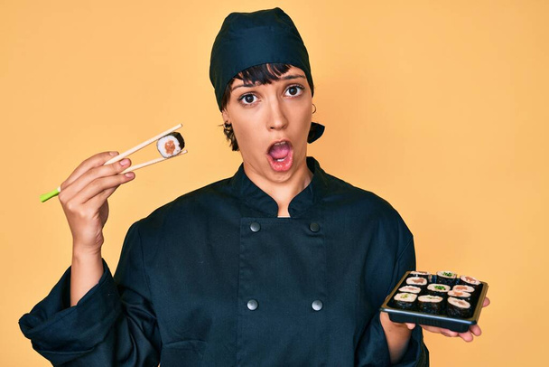 Mooie brunettte vrouw chef-kok met sushi met behulp van stokjes in shock gezicht, op zoek sceptisch en sarcastisch, verrast met open mond  - Foto, afbeelding