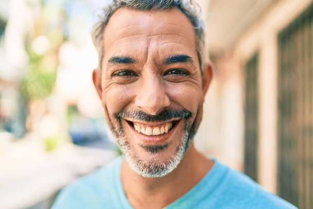 Hombre de pelo gris de mediana edad sonriendo feliz caminando en la calle de la ciudad. - Foto, Imagen
