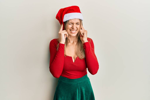 Όμορφη καυκάσια γυναίκα φορώντας χριστουγεννιάτικη φορεσιά και καπέλο που καλύπτει τα αυτιά με τα δάχτυλα με ενοχλημένη έκφραση για το θόρυβο της δυνατής μουσικής. κωφή έννοια.  - Φωτογραφία, εικόνα