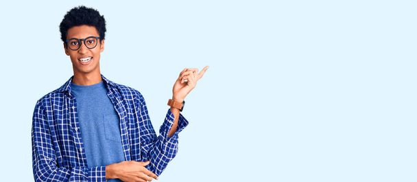 Jeune homme afro-américain portant des vêtements décontractés et des lunettes avec un grand sourire sur le visage, pointant avec la main et le doigt vers le côté regardant la caméra.  - Photo, image