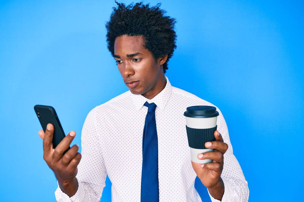 Hombre afroamericano guapo con cabello afro usando teléfono inteligente y bebiendo una taza de café relajado con expresión seria en la cara. simple y natural mirando a la cámara.  - Foto, Imagen