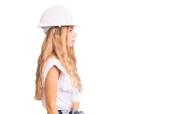 Beautiful caucasian woman with blonde hair wearing hardhat and painter clothes looking to side, relax profile pose with natural face with confident smile.  - Foto, Bild