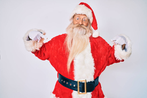 Γέρος ηλικιωμένος άνδρας με γκρίζα μαλλιά και μακριά γενειάδα φορώντας κοστούμι Άγιος Βασίλης αναζητούν αυτοπεποίθηση με χαμόγελο στο πρόσωπο, δείχνοντας τον εαυτό του με τα δάχτυλα περήφανος και χαρούμενος.  - Φωτογραφία, εικόνα